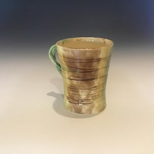 May include: A handmade ceramic mug with a cylindrical shape. The mug features a light brown and tan color scheme with horizontal brown stripes. It has a green handle and is suitable for hot or cold beverages.