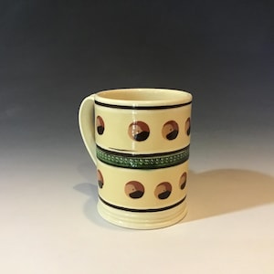May include: A cream-colored ceramic mug with a handle. The mug features two rows of brown and black circular designs. A green and black patterned band encircles the middle. The mug has a simple, elegant design.
