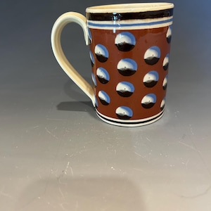 May include: A ceramic mug with a brown body decorated with white and blue polka dots. The rim has a dark brown band with blue and cream stripes. The handle is cream-colored.