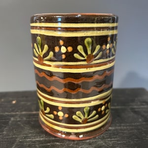 May include: A cylindrical ceramic mug with a dark brown base color. The mug is decorated with horizontal bands of yellow, orange, and brown, and floral patterns in green and brown. The mug has a glossy finish and a red rim.