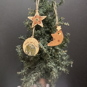 May include: A small Christmas tree decorated with three handmade ornaments. The ornaments include a star, a crescent moon, and a round disc with the words "Peace on Earth". The ornaments are brown and beige with twine hangers.