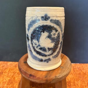 May include: A cylindrical ceramic mug with a cream-colored base and a dark blue decorative medallion. The medallion features an embossed white bird figure surrounded by small floral motifs. The mug has a smooth, slightly textured surface finish.