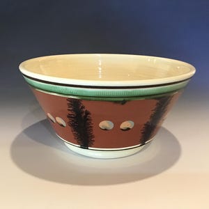 May include: A ceramic bowl with a cream-colored interior and a reddish-brown exterior. The bowl features a green and black band around the rim, with a design of black tree-like shapes and small, light-colored circles. The bowl is on a white surface.