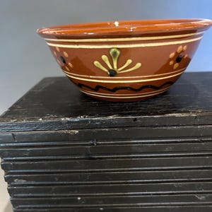 May include: A small, round, terracotta-colored bowl with decorative painted details. The bowl features a band of cream-colored paint with black and green floral designs. The bowl is set on a black, layered surface.