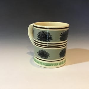 May include: A ceramic mug with a cream-colored interior and handle. The exterior features a light gray background with dark blue floral designs and brown stripes. The base has a green and white patterned border.