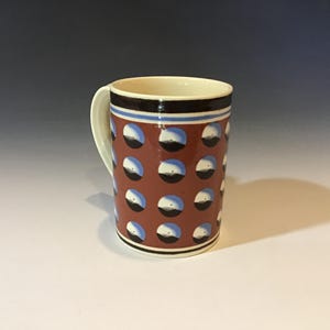 May include: A ceramic mug with a brown background and a repeating pattern of white, black, and blue circles. The mug has a white handle and a cream-colored interior. Black and blue stripes adorn the top.