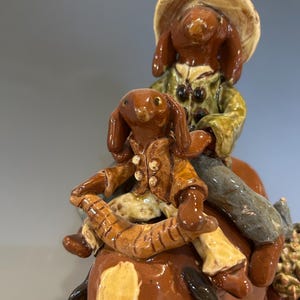 May include: A ceramic figurine depicting two dog-like figures riding a horse. The top figure wears a hat and green jacket, while the lower figure wears a brown jacket. The horse is brown and white.