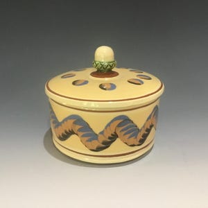 May include: A round ceramic lidded jar with a cream-colored base. The body features a horizontal brown and blue wavy pattern. The lid has a small green acorn-shaped finial and a ring of blue circular accents around the top edge.