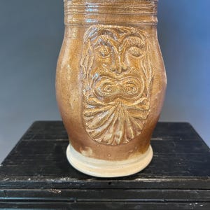 May include: A brown ceramic mug with a detailed face design. The mug has a cylindrical shape with a textured rim and a cream-colored base. The face is carved into the front of the mug, adding a unique, artistic touch to the pottery.