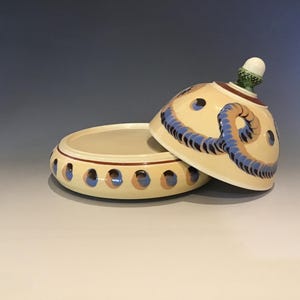 May include: A cream-colored ceramic trinket dish with a lid, featuring blue and brown decorative accents. The lid has a knob and a rope-like design. The dish has blue and brown accents around the rim. A decorative ceramic piece.