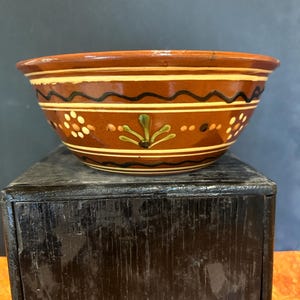 May include: A brown ceramic bowl with decorative painted bands. The bowl features a wavy black and white design, along with floral and dot patterns. The rim and base are a darker brown, creating a rustic, handcrafted aesthetic.