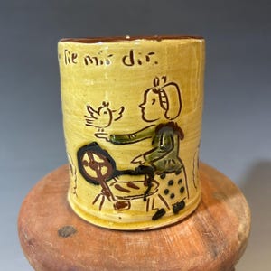 May include: A yellow ceramic mug featuring a brown hand-drawn illustration of a person spinning wool with a bird nearby. The text "lie mir dir" is inscribed near the top rim in brown lettering. The mug has a smooth, glossy finish and a cylindrical shape.