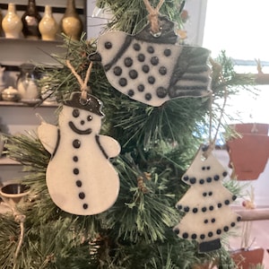 May include: Ceramic Christmas tree ornaments hanging on a green Christmas tree. The ornaments include a snowman, a bird, and a Christmas tree shape. Each ornament is white with black detailing and a twine hanger.