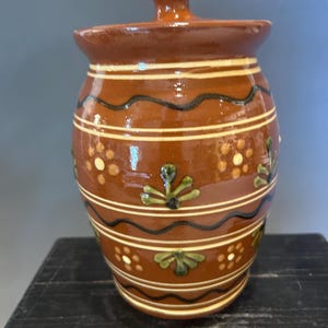 May include: A brown ceramic jar with a lid, decorated with horizontal white and black wavy lines, and green and white floral patterns. The jar has a rounded body and a small knob on the lid. The jar is sitting on a black surface.