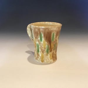 May include: A handmade ceramic mug with a handle. The mug features a beige base with brown, green, and yellow vertical streaks. The mug is suitable for hot or cold beverages.