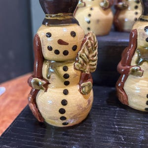 May include: A collection of ceramic snowman figurines. Each snowman has a brown top hat, a carrot nose, and black dot eyes and buttons. They are holding a leaf-shaped object and have brown arms.