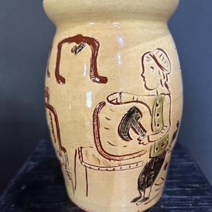 May include: A tan ceramic jar with a lid, featuring brown illustrations of people and objects. The jar has a rounded body and a lid with a brown knob. The illustrations depict figures in a rustic style.