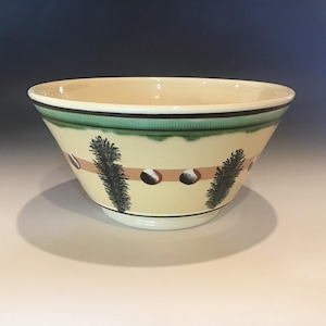 May include: A ceramic bowl with a cream-colored interior and exterior. The rim features a green band with black and white accents. The bowl is decorated with a brown stripe, dark green pine branches, and dark brown circular accents.