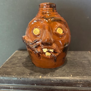 May include: A brown glazed ceramic face jug featuring a sculpted human-like visage with wide yellow eyes, a prominent nose, and an open mouth revealing three light-colored teeth. A small, dark, lizard-like creature is attached to the side of the face.