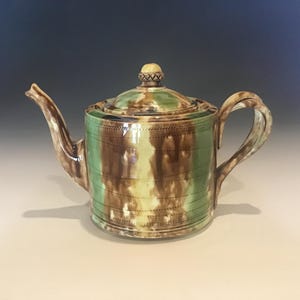 May include: A ceramic teapot with a lid, spout, and handle. The teapot is decorated with vertical stripes of green, brown, and yellow. The lid has a small, round knob. The teapot is likely used for brewing tea.