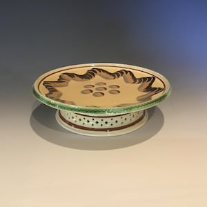 May include: A ceramic pedestal cake stand with a cream-colored top featuring brown and black decorative swirls and a green rim. The base is white with a pierced design and a brown accent.