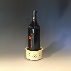 May include: A dark wine bottle with a label is displayed in a cream-colored, round wine bottle holder. The holder has a lattice design. The bottle is centered and stands upright.