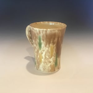 May include: A handmade ceramic mug with a handle. The mug features a unique design with brown, yellow, and green accents. The mug is approximately 5 inches tall.