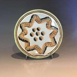 May include: A decorative ceramic plate with a light blue center and a star-shaped design. The star is formed by a series of interconnected, curved shapes in shades of tan and gray. The plate has a green border and is displayed on a metal stand.