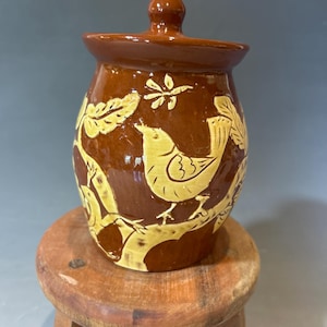 May include: A brown ceramic jar with a matching lid, featuring a glossy finish and light yellow sgraffito-style carvings of birds perched on branches surrounded by leaves. The jar has a rounded body and a small knob handle on the lid.