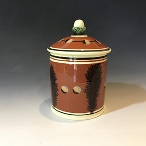 May include: A ceramic jar with a lid, featuring a reddish-brown base color. The jar is decorated with black tree-like designs and circular cutouts. The lid has a white and black trim, with a green and white knob on top.