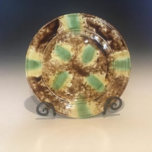 May include: Handmade ceramic plate with a unique design. The plate features a brown, green, and beige speckled pattern. The rim has alternating green and beige stripes. The plate is displayed on a decorative metal stand.