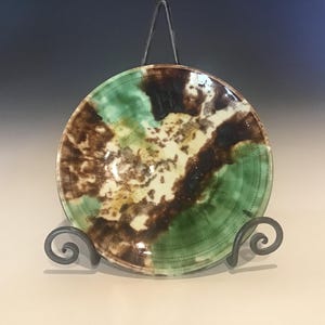 May include: A handcrafted ceramic plate with a unique design. The plate features a blend of green, brown, and cream colors. It is displayed on a decorative metal stand, showcasing its artistic appeal. The plate's diameter is approximately 10 inches.