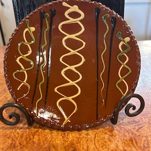 May include: A round, brown ceramic plate with a decorative design. The plate features a series of vertical lines and looping patterns in cream, black, and green. The plate is displayed on a decorative metal stand.