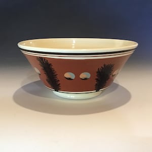 May include: A ceramic bowl with a reddish-brown exterior, featuring black tree-like designs and circular accents. The rim has black and white stripes. The interior is cream-colored, and the bowl sits on a white base.