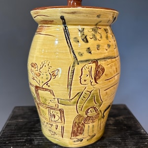 May include: A ceramic jar with a lid, featuring a hand-painted design. The jar is primarily yellow with brown and black accents. The artwork depicts a person with a basket and a rooster. The lid has a brown knob.
