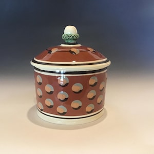 May include: A ceramic jar with a lid, featuring a reddish-brown base and lid adorned with light blue and orange polka dots. The jar has a white rim and a decorative knob on top, with a green and white design.