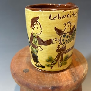 May include: A yellow ceramic mug with a brown rim and handle, featuring a hand-painted illustration of a person standing near a bird perched on a fence. The text "Lebenicht" is written in brown script near the top edge of the mug's exterior surface.