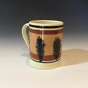 May include: A ceramic mug with a cream-colored handle and base. The mug features a design of alternating bands of tan, brown, and black, with black tree-like shapes. The mug is approximately 4 inches tall.