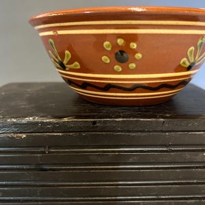 May include: A ceramic bowl with a reddish-brown exterior, adorned with cream-colored horizontal stripes and floral designs in green and black. The bowl's interior is a lighter shade of brown.