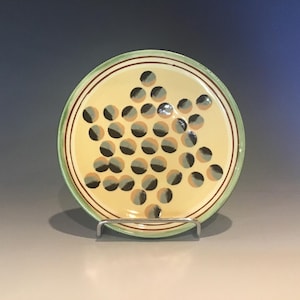 May include: A round ceramic plate with a light yellow center, decorated with black and tan dots. The rim has a green border with brown accent lines. The plate is displayed on a silver metal stand.