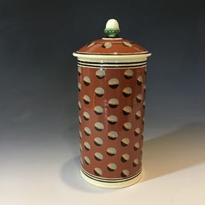 May include: A tall, cylindrical ceramic jar with a lid. The jar is a reddish-brown color with a pattern of evenly spaced, light blue and black polka dots. The lid is the same color as the jar, with a white rim and a small, white knob on top.