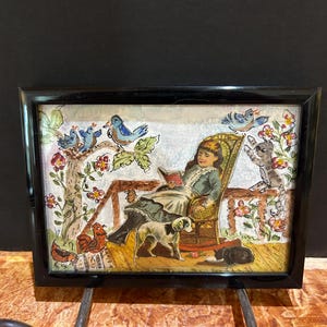 May include: A framed illustration depicts a person reading in a rocking chair, surrounded by birds, a cat, and dogs. The artwork features a black frame and a warm color palette with floral accents. The scene evokes a cozy, pastoral theme.