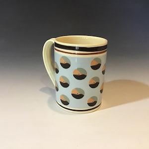 May include: A ceramic mug with a light blue background and a repeating pattern of black, tan, and teal half-circles. The mug has a cream-colored interior and handle, with brown and black bands near the rim.