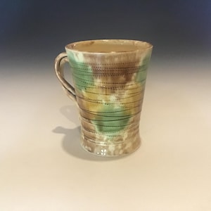 May include: A handmade ceramic mug with a handle. The mug features a color scheme of brown, green, and yellow, with horizontal black dotted lines. The mug is suitable for hot or cold beverages.