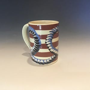 May include: A ceramic mug with a white handle and a cylindrical body. The body features alternating red and white horizontal stripes. A blue and black design wraps around the mug. The mug is suitable for hot or cold beverages.