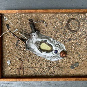 May include: A silver and copper sculpture of a creature with a single eye, set in a wooden frame with a sand-like background.