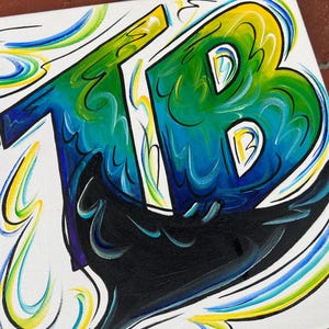 May include: A square canvas painting featuring the letters "TB" in vibrant colors. The letters are primarily blue and green, with yellow accents and black outlines. Swirls of blue, green, and yellow surround the letters against a white background.