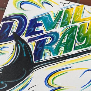 May include: A canvas painting featuring the words "DEVIL RAYS" in large, colorful letters. The letters are shaded in blue, green, and yellow, with black outlines. A black shape resembling a ray's wing is in the foreground, with blue and yellow swirls.