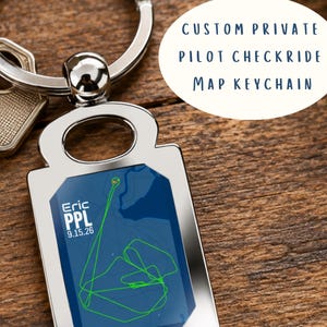 May include: A rectangular silver-toned metal keyring featuring a dark blue map insert with a bright green flight path graphic. The text reads Eric, PPL, and 9.15.26. A silver metal ring is attached to the top of the keyring, next to a metal key.