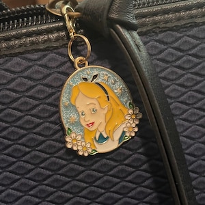 May include: A gold-coloured metal charm depicting Alice from Alice in Wonderland. The oval charm has a blue background with stars and white flowers. Alice has blonde hair and wears a blue dress. The charm is attached to a black bag.